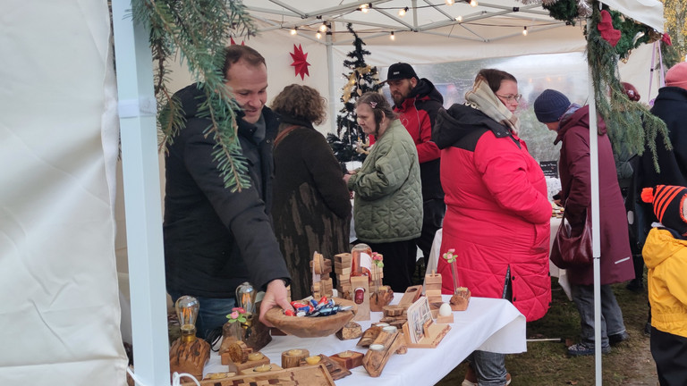 Mehrere Personen in bunten jacken stehen an einem Stand mit Holzarbeiten auf dem Weihnachtsmarkt des Immanuel Haus Ecktannen Mehrere Personen in bunten jacken stehen an einem Stand mit Holzarbeiten auf dem Weihnachtsmarkt des Immanuel Haus Ecktannen