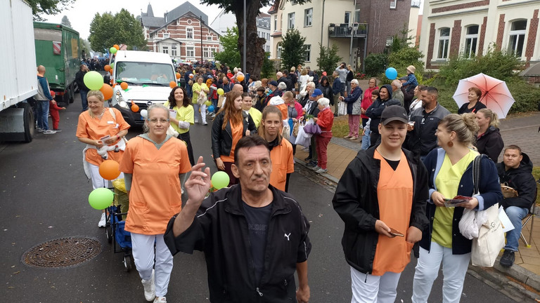 Mitarbeitende auch in Funktionskleidung laufen vor dem Transporter beim Festumzug zum Jubiläums-Müritzfest mit Pflegekräften, Bewohnerinnen und Bewohnern aus dem Immanuel Haus Ecktannen 