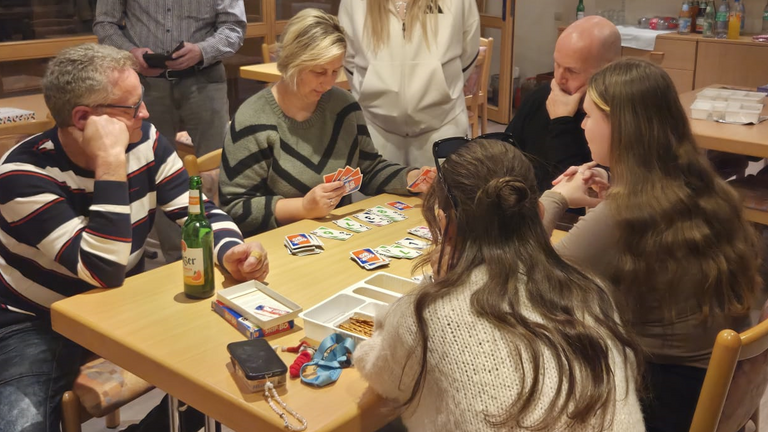 Mehrere Teammitglieder sitzen und stehen an einem Tisch beim Kartenspielen in der Immanuel Begegnungsstätte Ecktannen im Heilbad Waren beim gemeinsamen Spieleabend für Mitarbeitende der Seniorenpflegeeinrichtung mit Tagespflegeangebot Immanuel Haus Ecktannen.